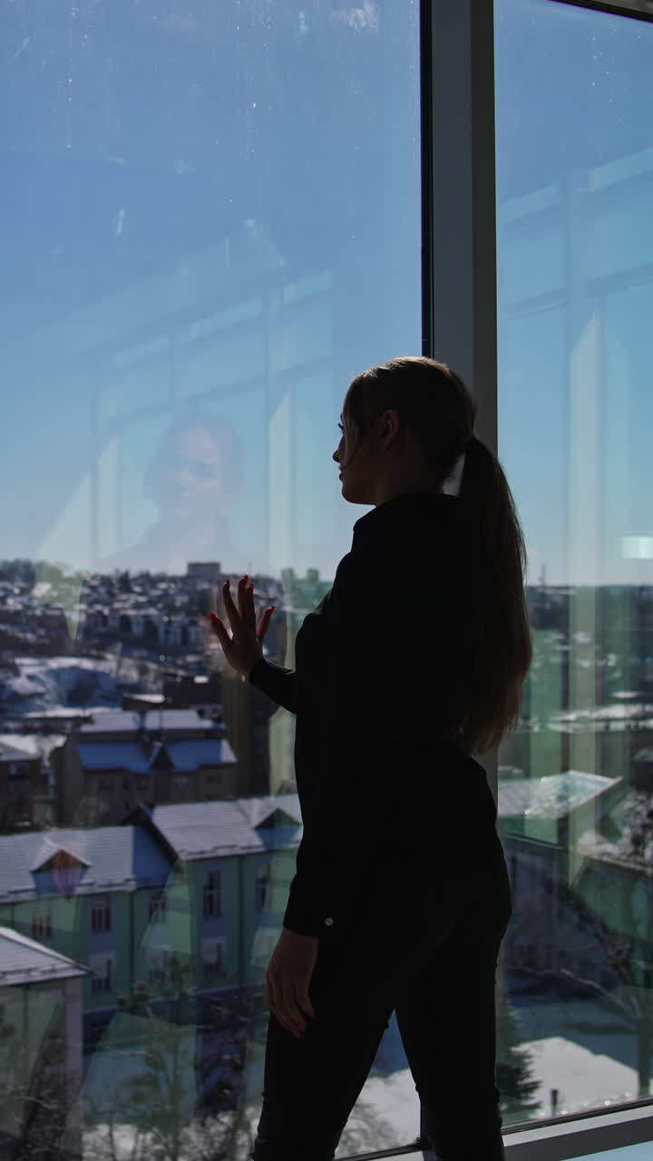 Young good-looking female wearing dark clothes standing in front of the panoramic windows. Sunrays coming into the big office room. Girl looking out of the window and enjoying the cityscape. Vertical video