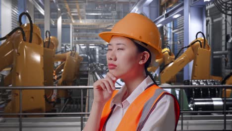 close up side view of asian female engineer with safety helmet thinking about something and looking around while standing with industrial robotic arms working