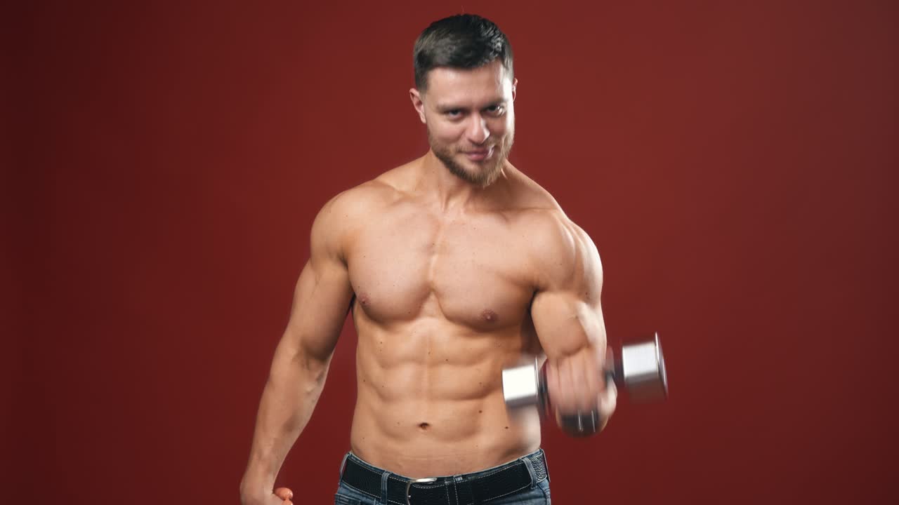 Muscular bodybuilder on dark red background. Athlete with naked torso lifting sausage in one hand and dumbbell in other. Smiling sportsman in studio.