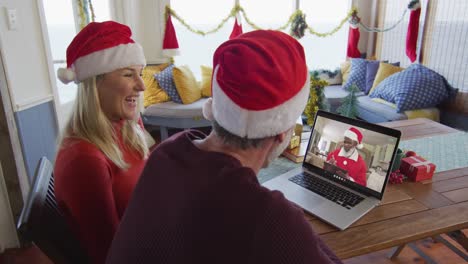 Smiling-caucasian-couple-with-santa-hats-using-laptop-for-christmas-video-call-with-santa-on-screen