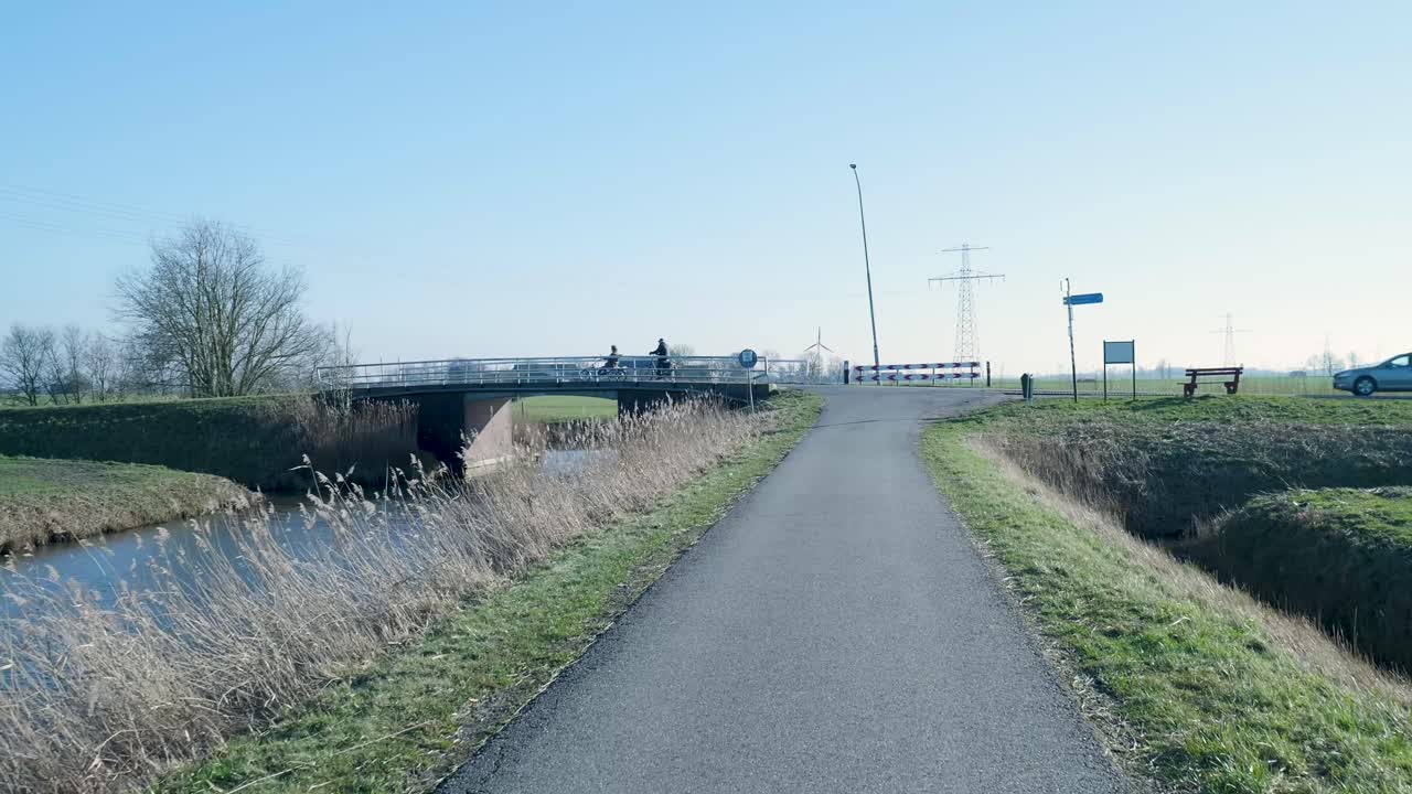 Cyklister og bil krydser en bro over en kanal i landligt landskab