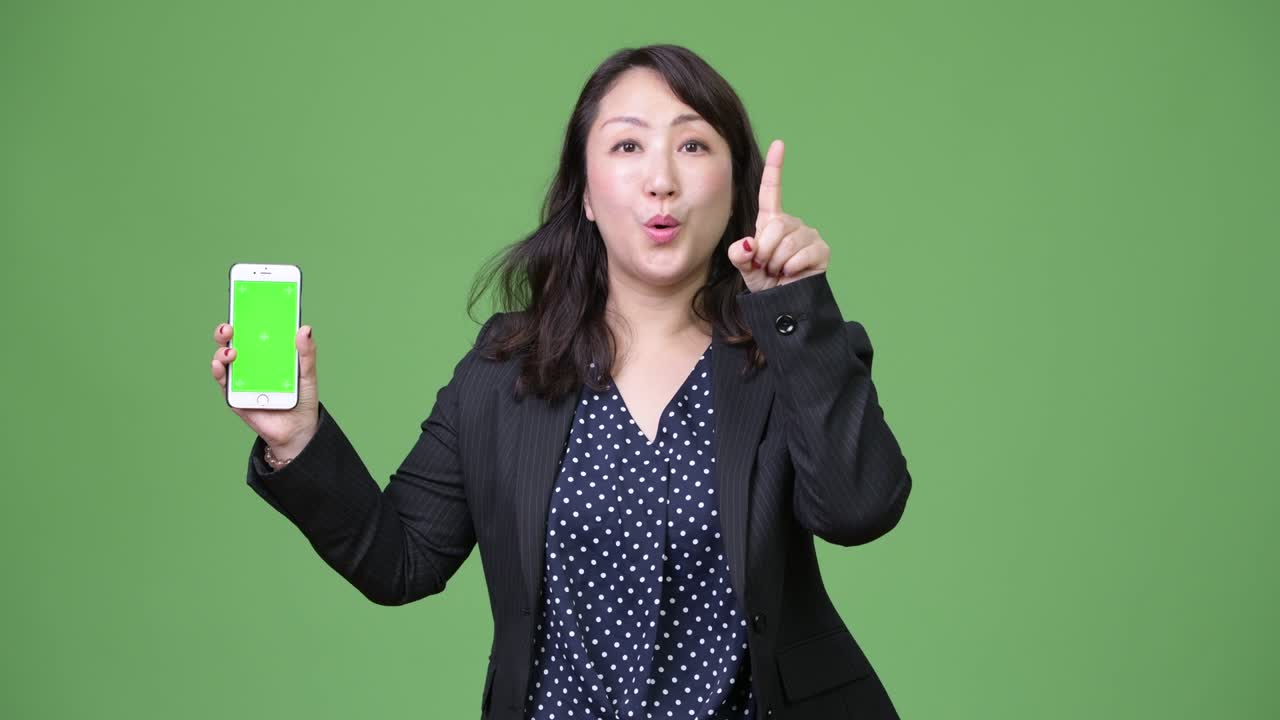 Mature beautiful Asian businesswoman showing phone while pointing finger up