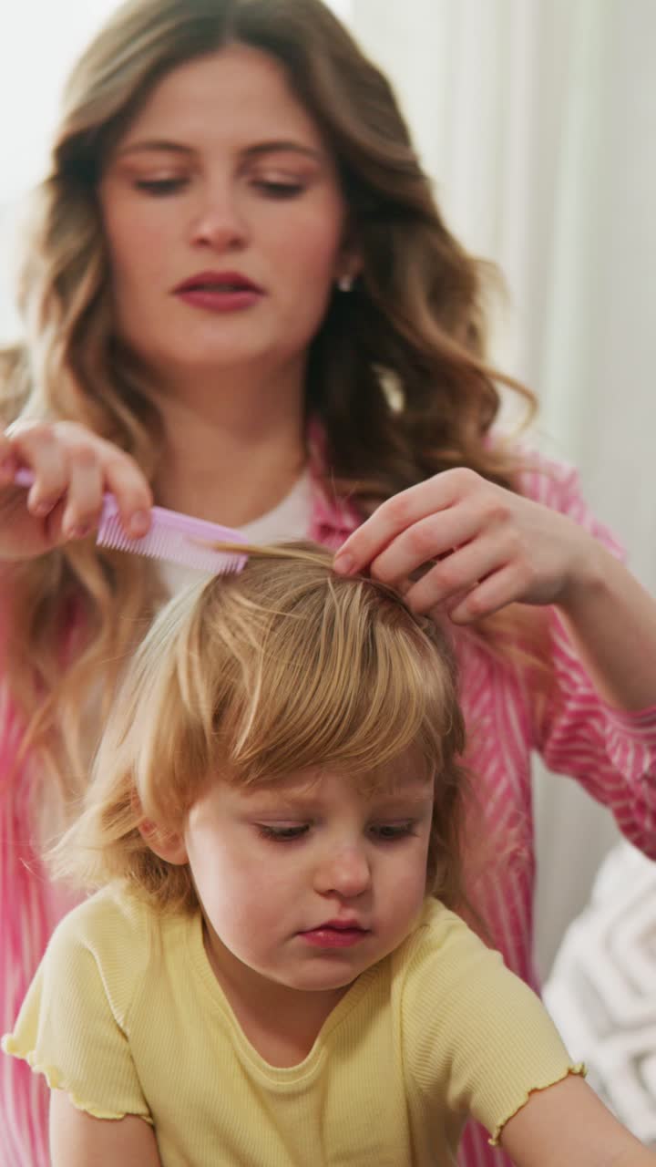 Mother gently brushes her toddler's hair. Child looks down, focused on hands. Scene shows quiet, caring routine. Ideal for parenting, home life, or beauty content