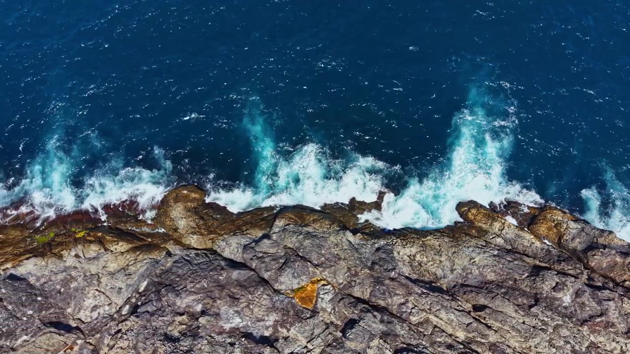 Golven slaan tegen rotsachtige kust onder helderblauwe lucht tijdens de middag op een kustlocatie.