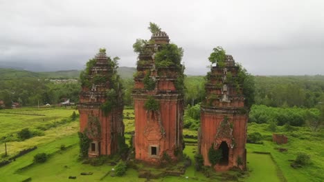 aerial view of three ancient red brick cham towers overgrown with vegetation, surrounded by lush green fields and trees