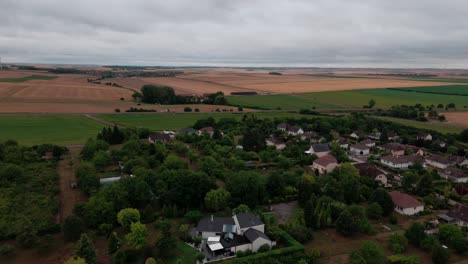 aerial drone shot descending over a charming french village surrounded by vast agricultural fields. shows houses with gardens and pools amidst lush trees under a cloudy sky