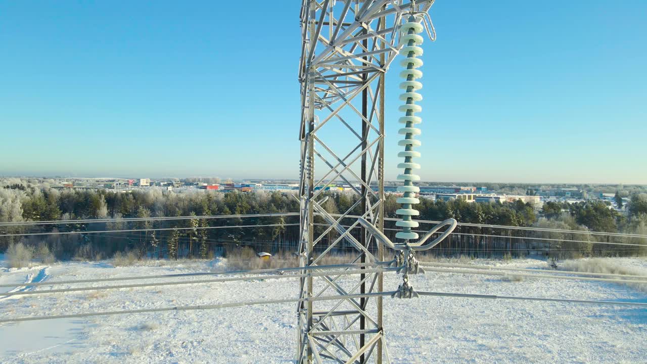 Close aerial movement reveals frozen power line insulators and icy cables, descending beside the pylon while snow covered fields, forest, and distant city form a crisp winter backdrop