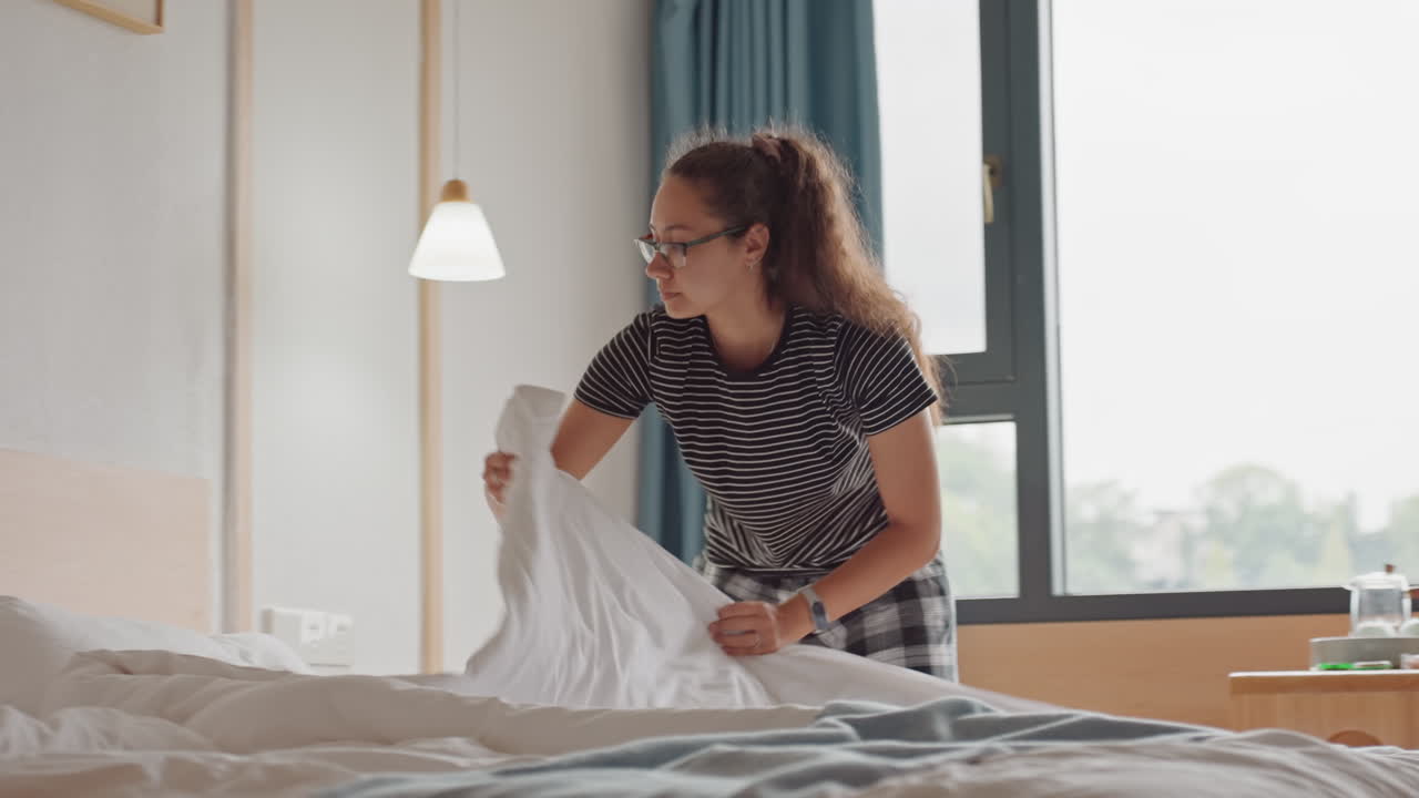 Mujer haciendo la cama del hotel en una habitación moderna y luminosa, ajustando sábanas y almohadas, luz solar a través de la ventana, cortinas azules, lámpara de noche, enfocada en la pulcritud, viajera solitaria preparando el espacio para su estancia.