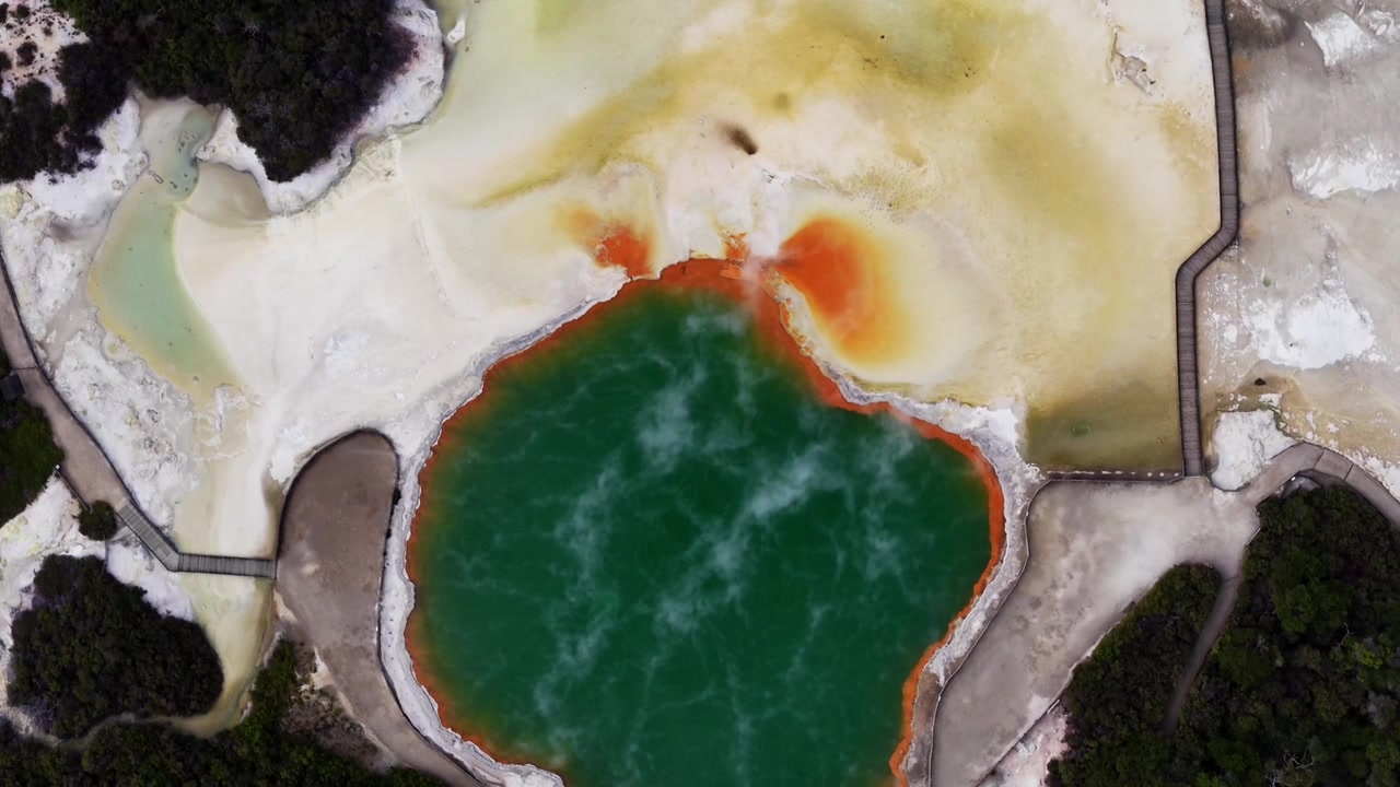 Pohled dronem shora na geotermální terén Rotoruy, odhalující hluboké smaragdové termální jezírko ohraničené oranžovými minerálními usazeninami, bledou zemí a tmavou původní vegetací.
