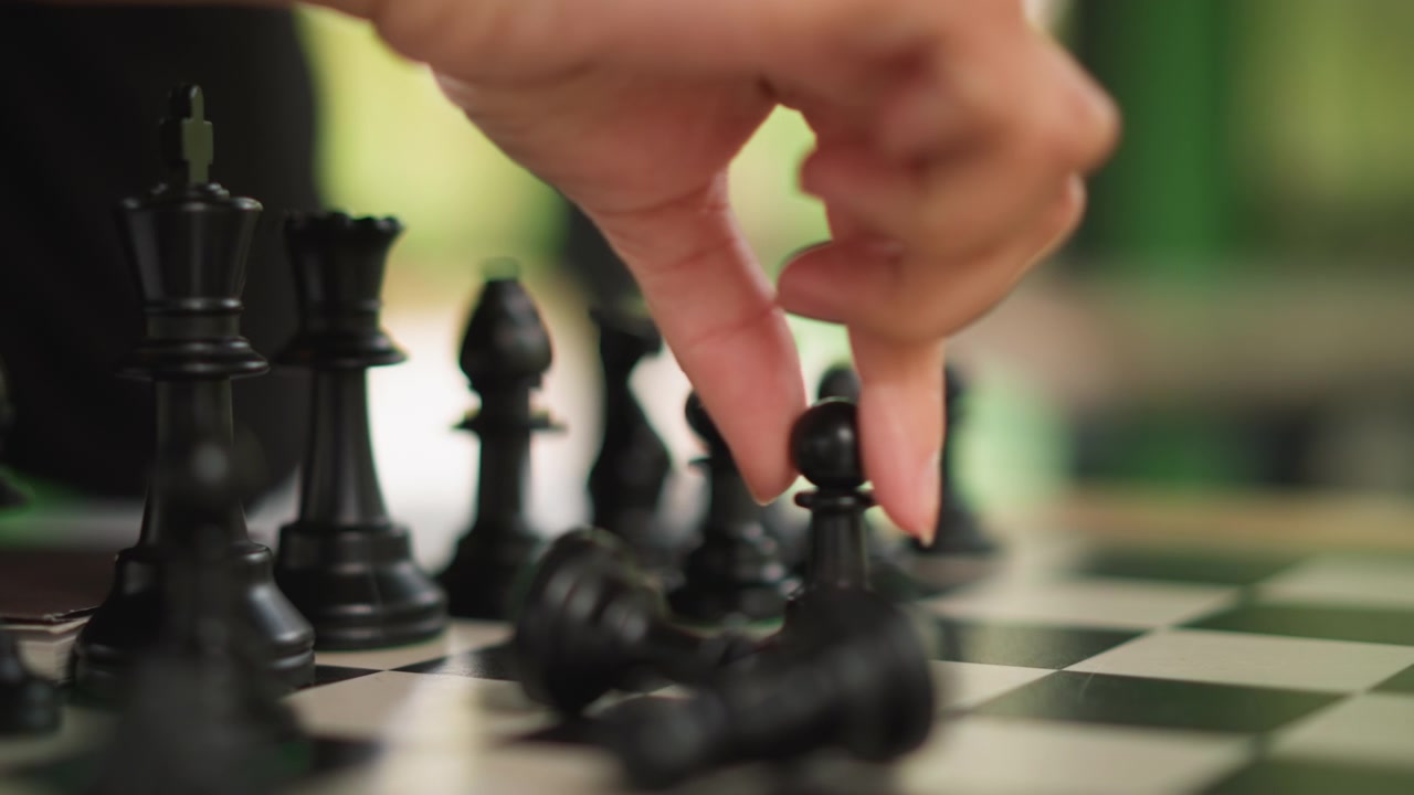 Primo piano di mano che muove pezzi degli scacchi neri su una scacchiera durante il gioco. La messa a fuoco rimane stretta per tutta la scena, catturando l'interazione tra dita e pedoni mentre altri elementi del gioco sono sfocati