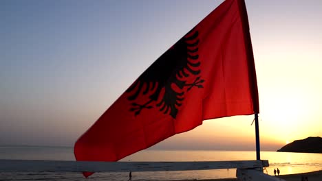 albanian flag fluttering in the wind, red with a black double-headed eagle in the center, with a sunset over the adriatic sea in the background