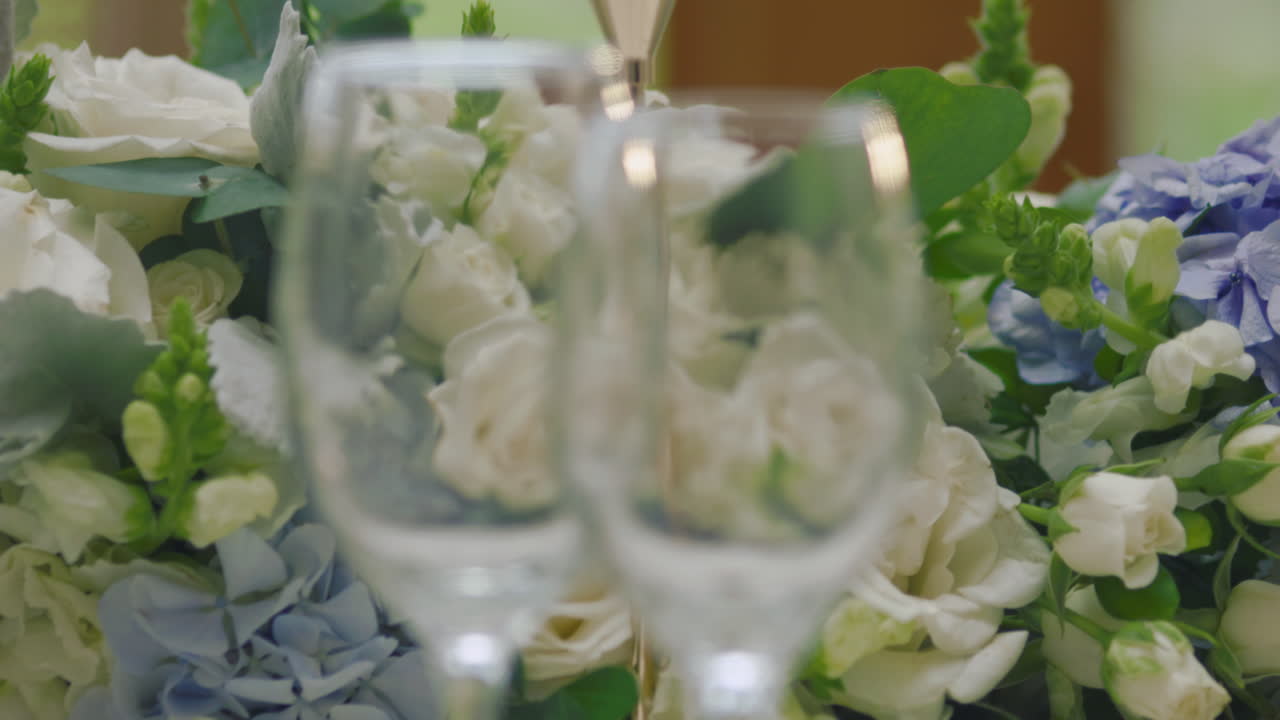 An elegant visual detail where the initial focus shows glass cups on the table and, through gentle panning, it blurs to focus on a delicate floral arrangement.
