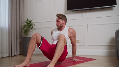 A-confident-athletic-man-with-stubble-in-a-white-T-shirt-and-red-shorts-does-special-exercises-to-develop-the-muscles-of-the-whole-body-during-his-morning-exercises-on-the-carpet-in-a-modern-bright-apartment