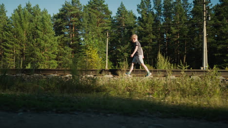 calm little boy in dark t-shirt and shorts walking alone near rail track surrounded by tall grass, gravel, and green forest with power lines and poles under bright daylight in quiet countryside scene
