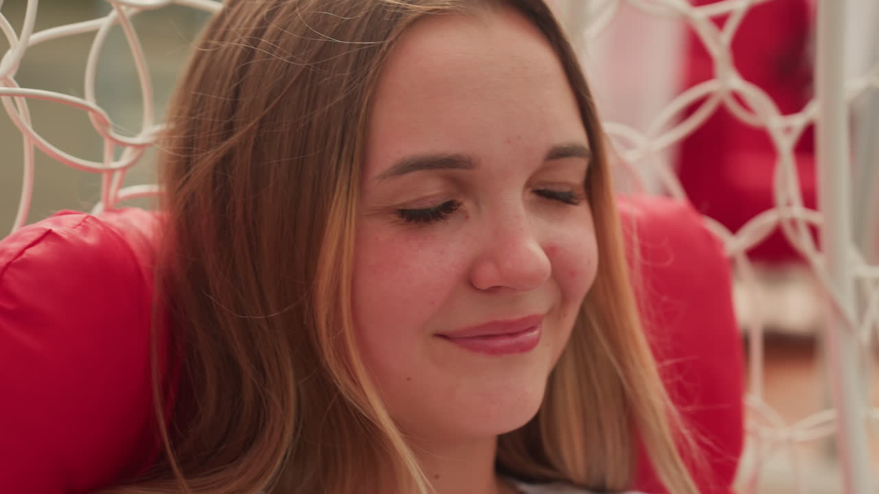 Giovane donna sorridente su una sedia sospesa, ritratto ravvicinato con espressione rilassata, lentiggini, cuscino rosso, sfondo con rete in macramè, luce solare calda, occhi chiusi, sorriso gentile, toni cinematografici tenui, intimo