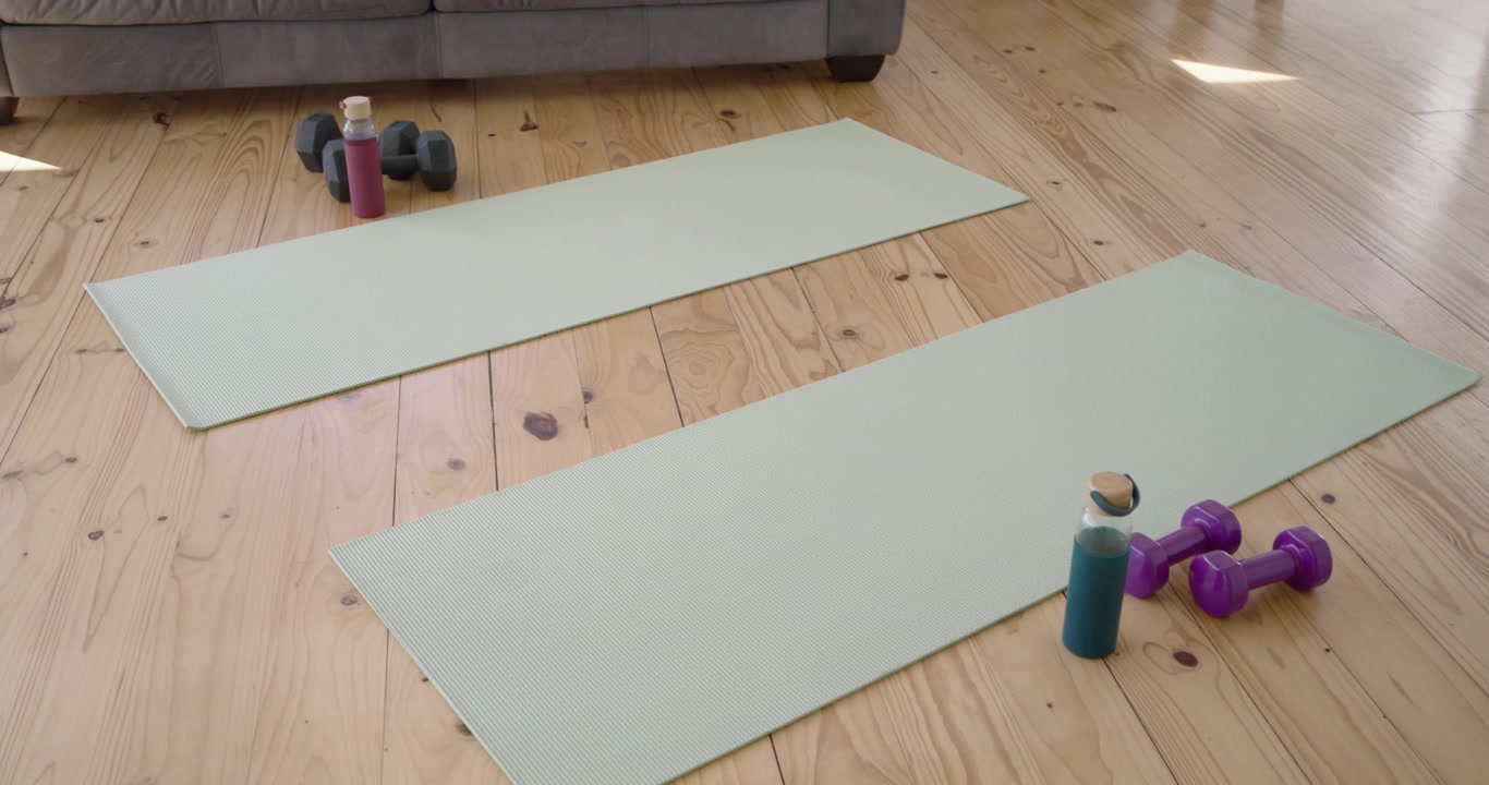 La cámara se aleja, revelando dos esterillas de yoga color menta, botellas y mancuernas, mostrando una configuración de entrenamiento en casa