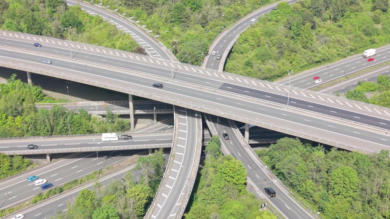 Aerial view of a massive motorway interchange with multiple layers of roads. Cars and trucks are actively moving in all directions during a bright day.