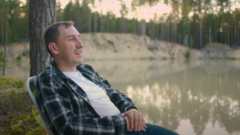 single man is relaxing at nature sitting in armchair on shore of forest lake and enjoying silence and calmness