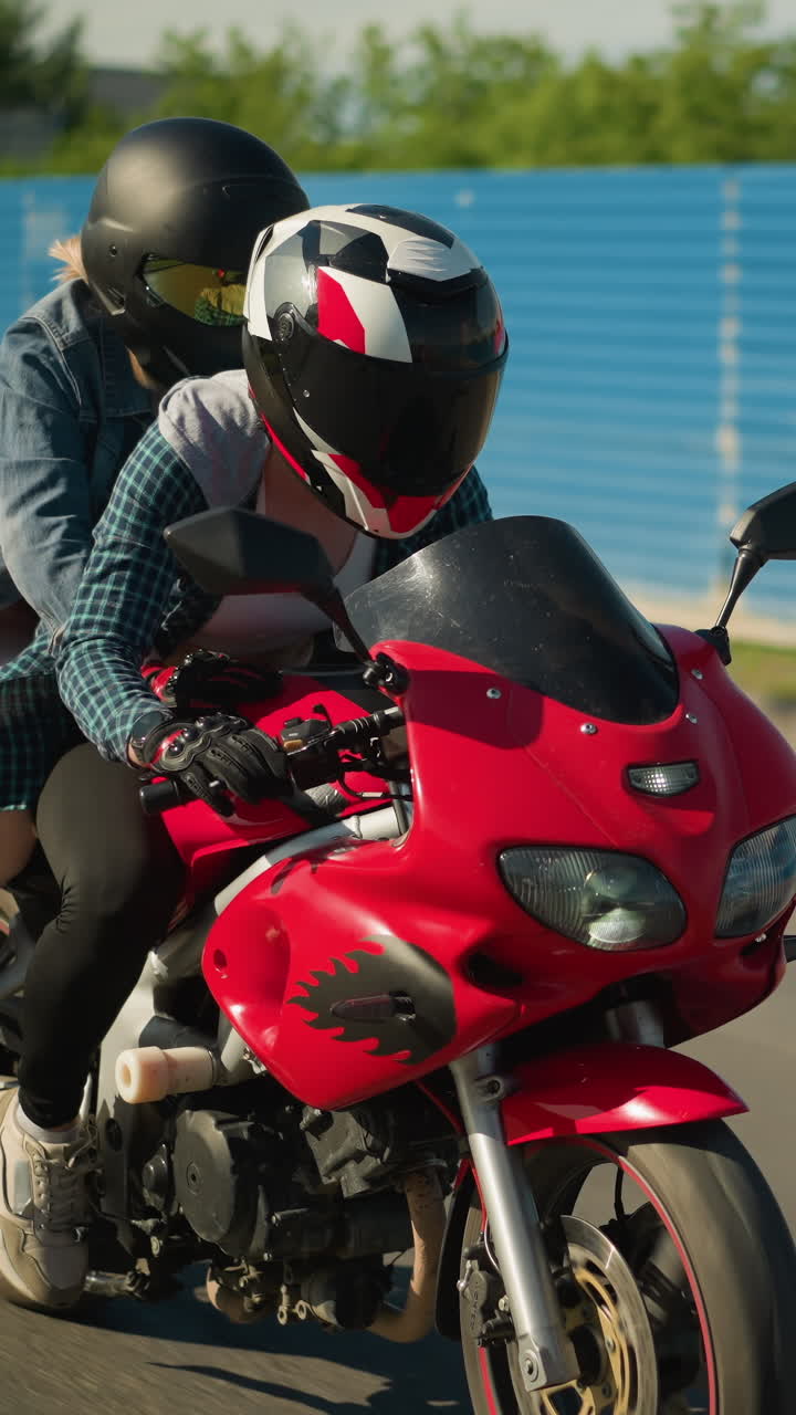 dos amigos viajan en una bicicleta eléctrica roja con cascos, pasando por un sitio de construcción con hojas de zinc azul, se ven coches con faros encendidos acercándose en el fondo cerca de un edificio de la ciudad