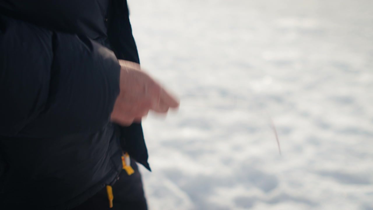 Tight side view of a bundled person slowly jigging a short rod over bright ice on a frozen lake surface.