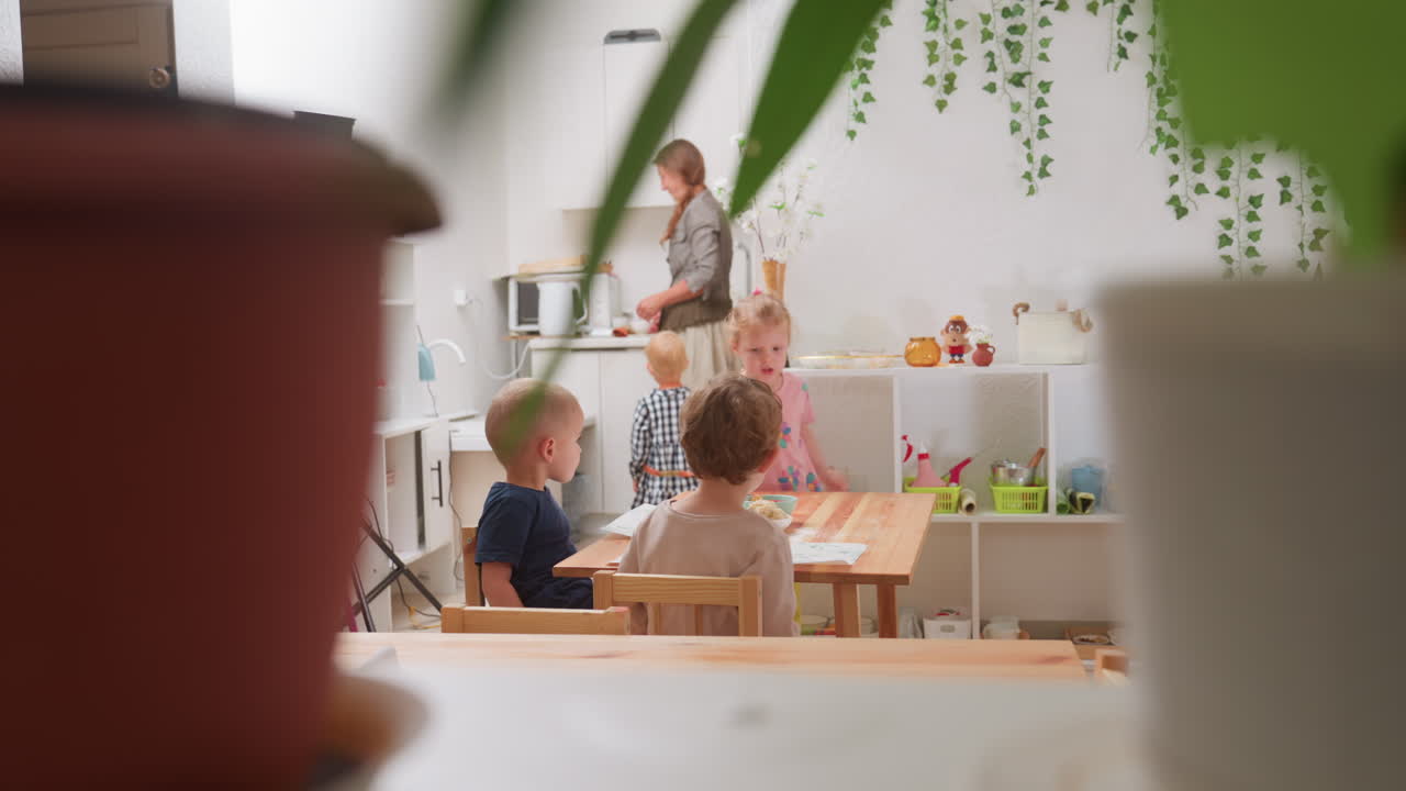 Scene in kindergarten viewed through decorative plants showing teacher near kitchen as girl in pink carries bowl with apple slices toward table while other children sit engaged