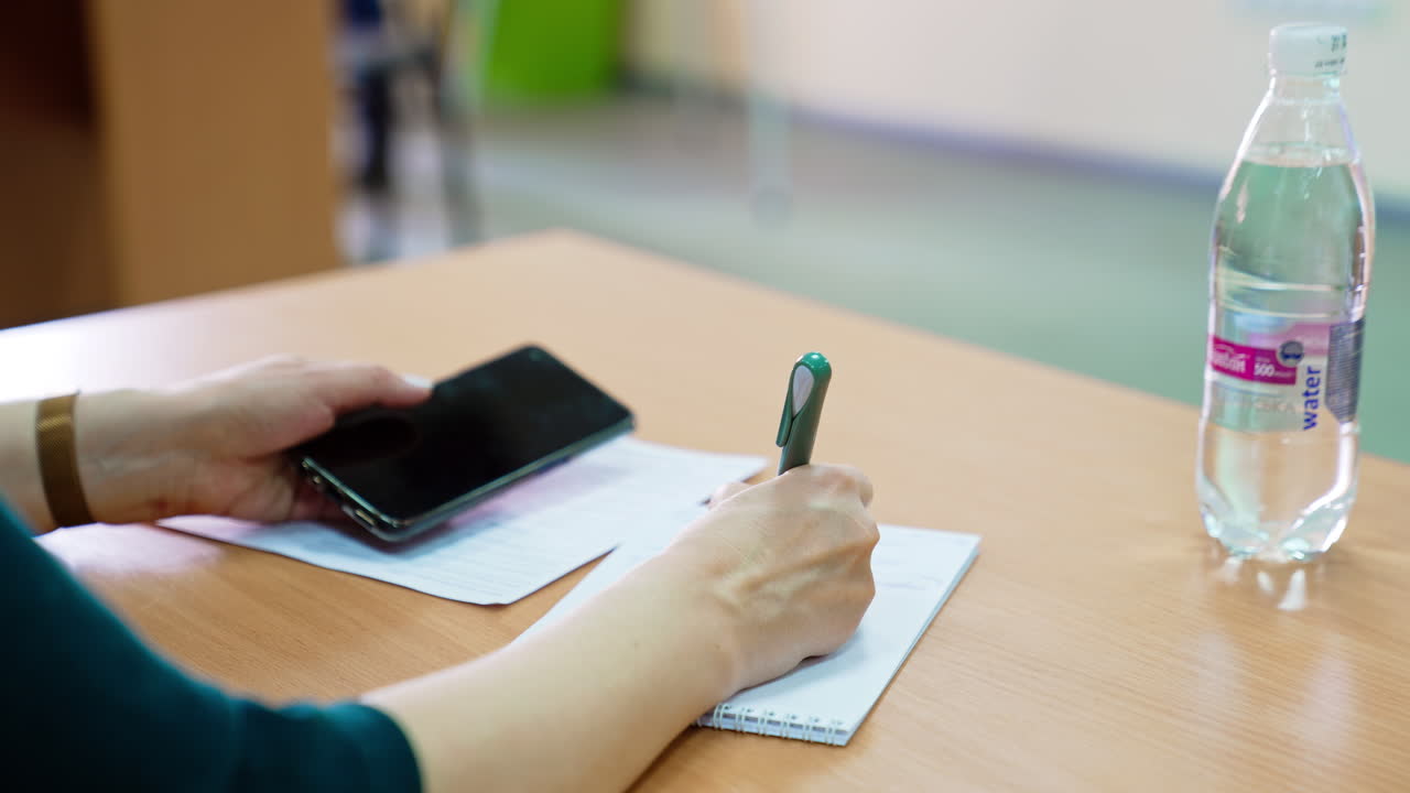 Unrecognized woman taking notes into paper notebook. Woman holding a phone in another hand. Blurred backdrop.