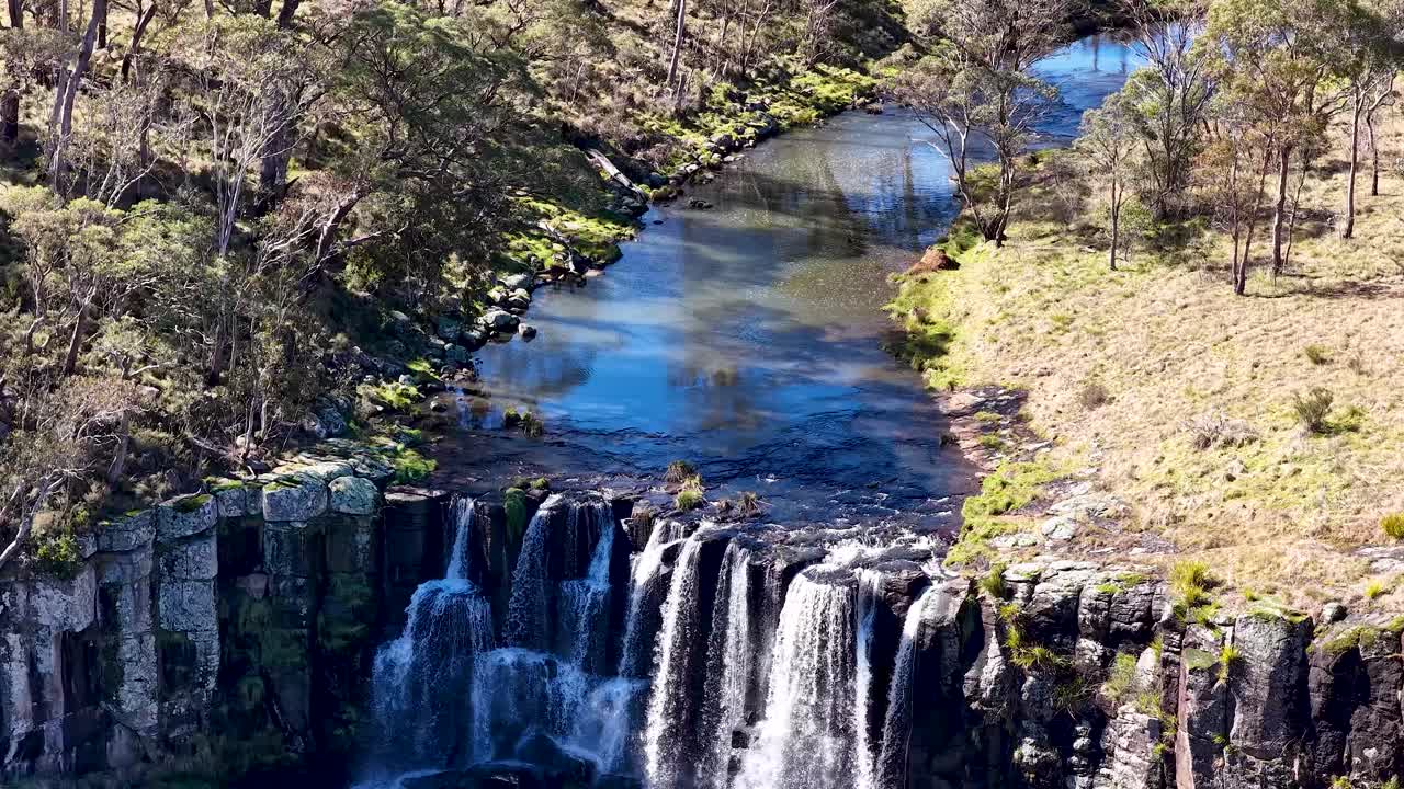 Drone footage captures Ebor Waterfall cascading over basalt cliffs into a lush gorge, surrounded by forest and grassland, under bright natural daylight