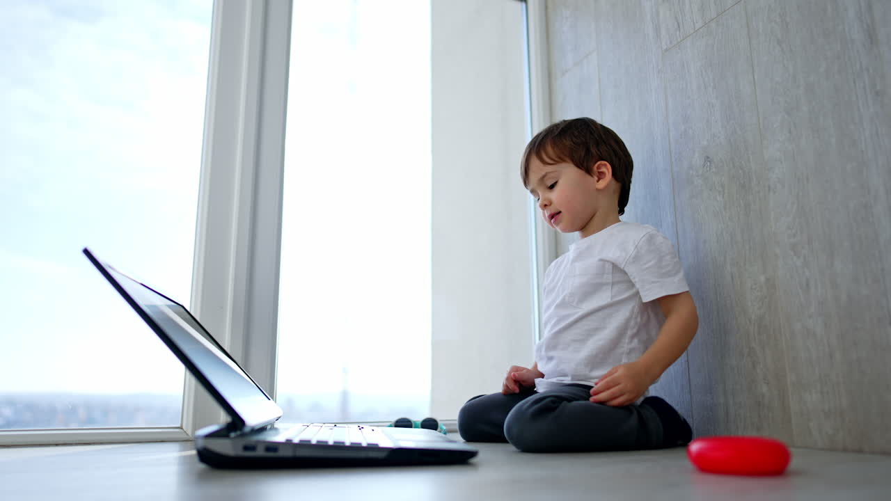 Little Caucasian baby boy sits on his knees. Cute child is focused on the laptop in front of him. Low angle view.