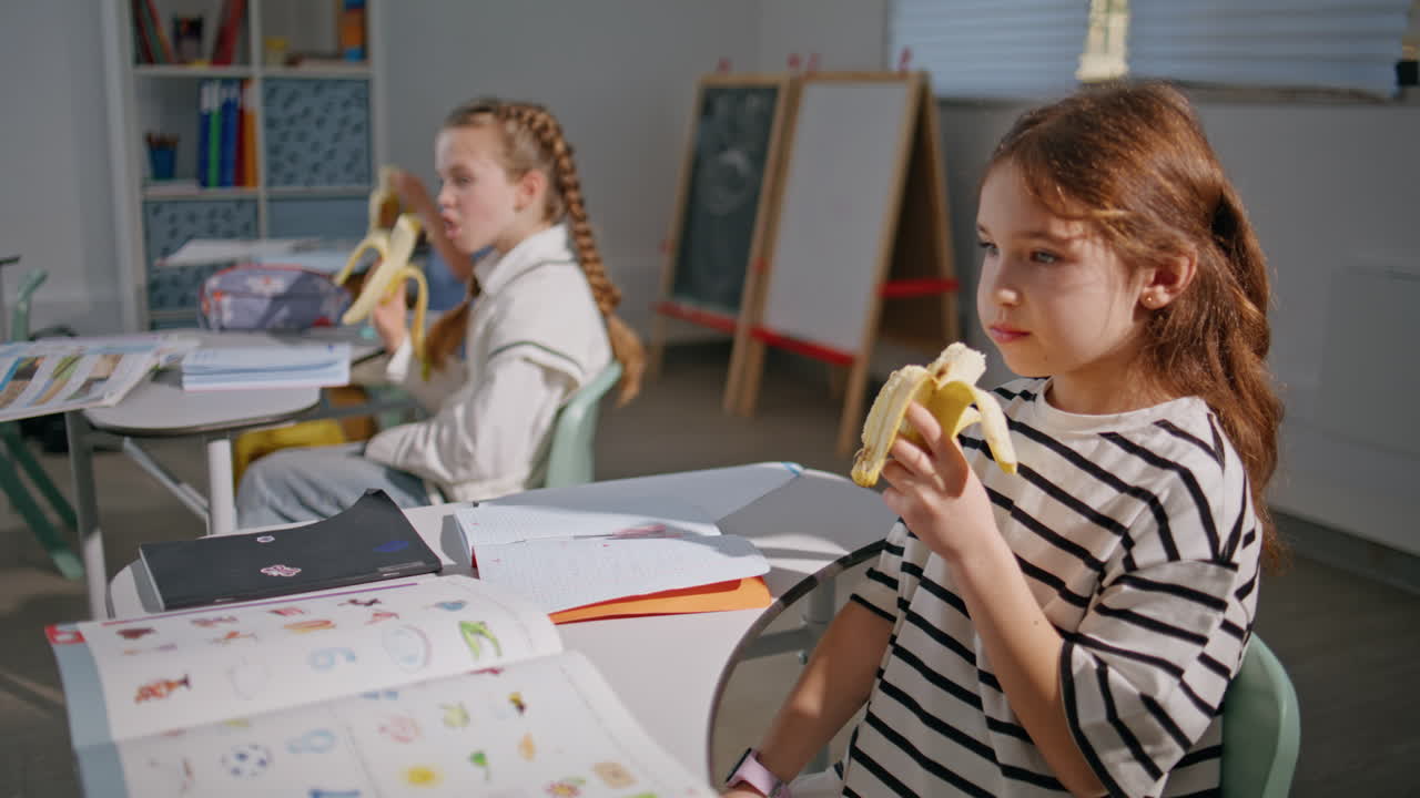 School kids eating bananas at desks closeup. Hungry classmates having lunch