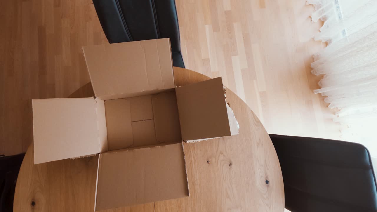 A man sets up an empty cardboard box on a wooden table, preparing for packing or shipping. Ideal for logistics, e-commerce, moving, packaging, or supply chain concepts
