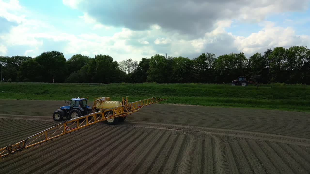 A tractor sprays the field with crop protection products on a partly cloudy day, near a road where another tractor is driving