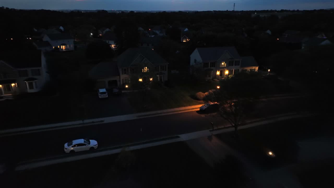 Aerial flight over dark american housing area with street and lighting window of House. Drone landing shot. Parking cars on street. Dusk scene in Suburbia.