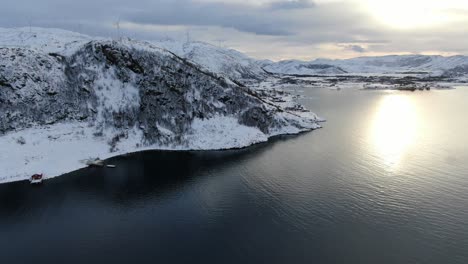 Drone-view-in-Tromso-area-in-winter-flying-over-a-snowy-mountain-peak-with-the-sea-in-front-and-wind-mills-on-the-mountains-in-Norway