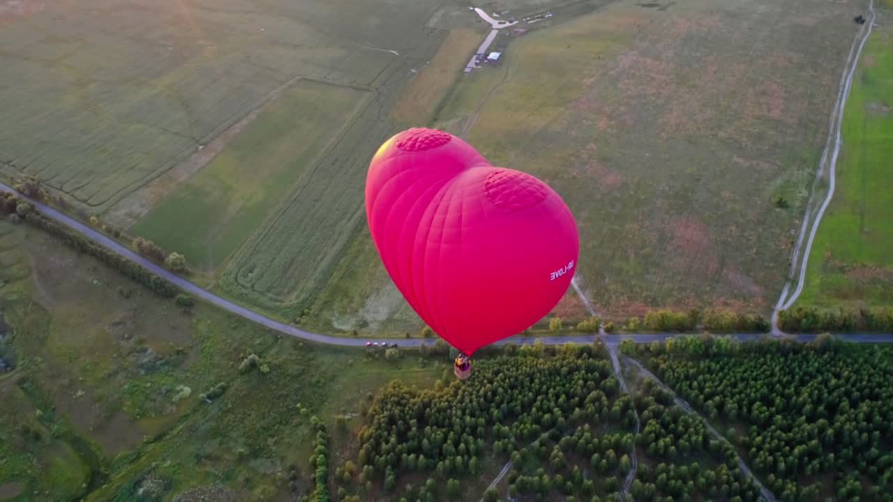 Red aerostat over fields. Romantic hot air balloon in a form of a heart travels in the sky on beautiful nature background at sunset. Top aerial view.
