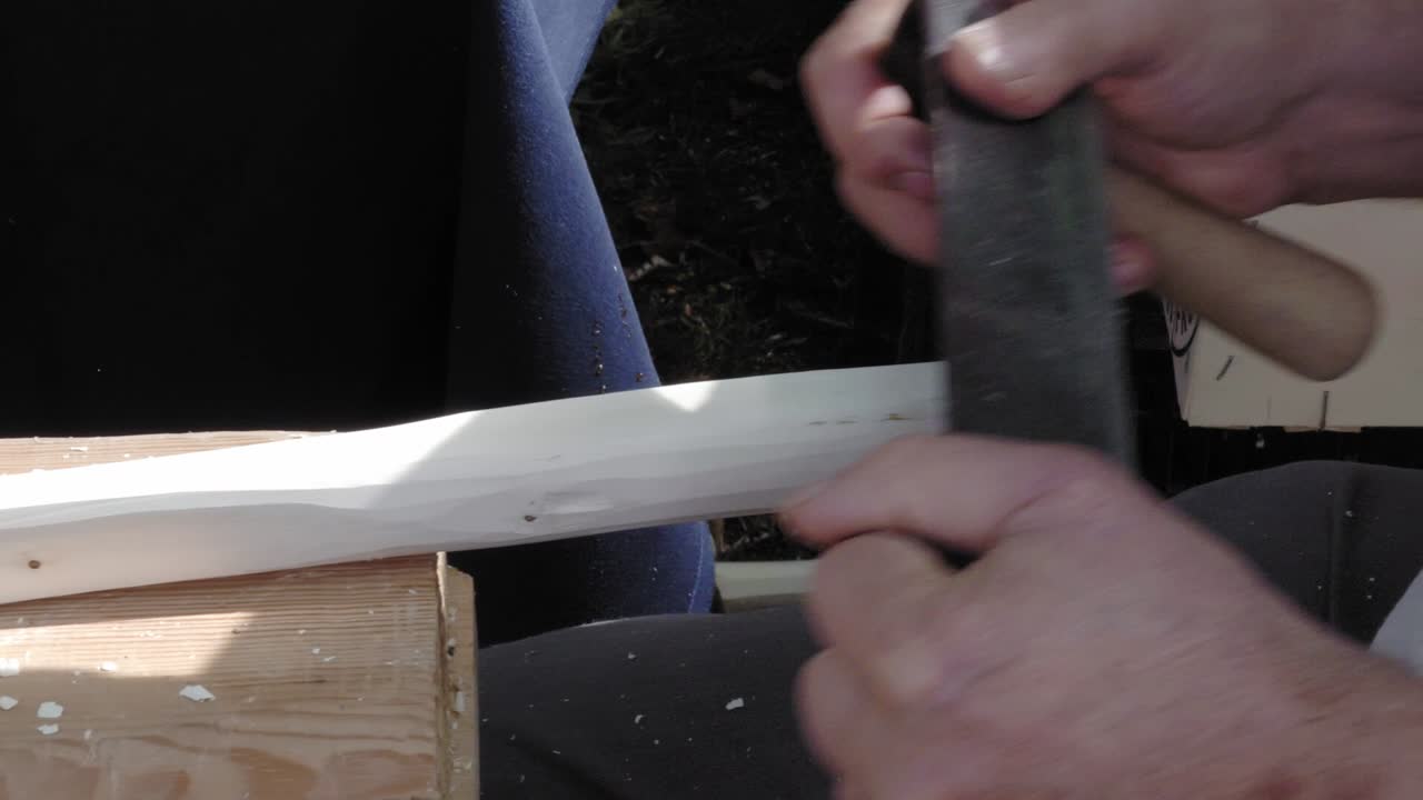 A traditional carpenter uses a metal planer to peel wood. Shavings are flying. He works outside during the event. People pass by. Close-up of hands. Slowmotion.