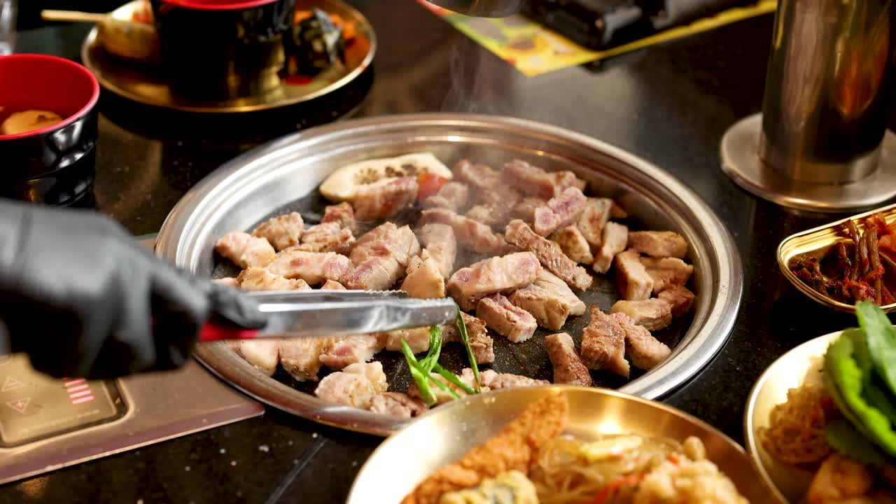 Gloved hand grilling pork belly pieces on tabletop grill, surrounded by side dishes, warm lighting