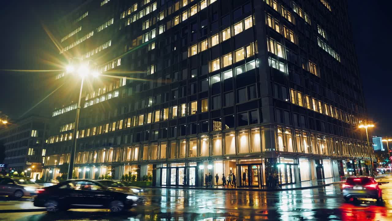 carretera urbana húmeda que refleja las luces brillantes de la ciudad, iluminando la fachada de un edificio contemporáneo con vehículos que pasan durante la hora pico nocturna