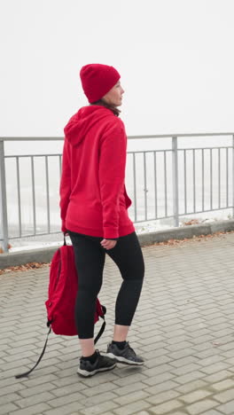 business woman holds backpack while rope drags along ground, walking on interlocked path beside iron railing in foggy atmosphere with dry scattered foliage and moving car visible below nearby bridge