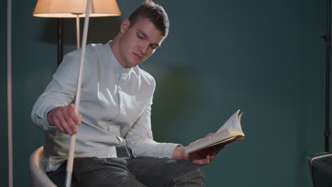 close-up of dude in white shirt and grey trousers sitting on white chair, reading book while holding cue stick. focused expression suggests deep concentration. warm lighting enhances thoughtful