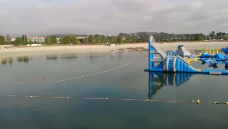 Schwimmendes-Schloss-Mit-Attraktionen-Und-Rutschen-Auf-Einem-Überwasser-Unterhaltungsrundgang-Mit-Booten,-Die-An-Einem-Bewölkten-Sommertag-Am-Strand-Schwimmen,-Panoramaaufnahme,-Die-Nach-Rechts-Rollt