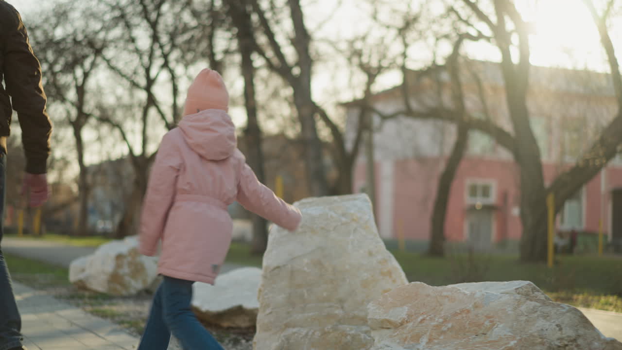 оживленная сцена с маленькой девочкой в розовой шапке и куртке, с энтузиазмом поднимающейся на небольшие скалы, в то время как мужчина в коричневой куртке следует за ним на заднем плане