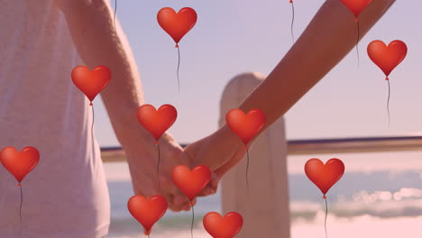 multiple heart balloons floating against mid section of couple holding hands at beach