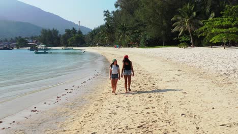 Stylish-beautiful-girls-walking-on-sand-of-exotic-beach-near-calm-clear-water-of-sea-and-tropical-trees-of-Ko-Pha-Ngan-island-in-Thailand