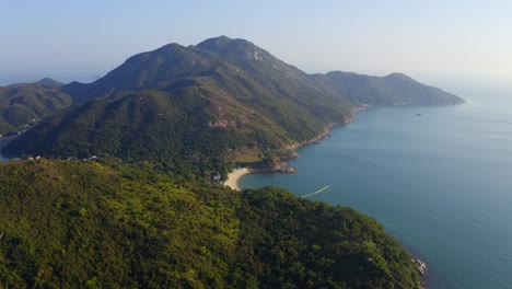 Cinematic-drone-shot-traveling-forward-and-panning-down-toward-a-beach-on-the-coast-of-a-tropical-island-with-green-mountains-on-it