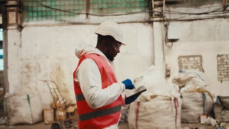 A-man-with-Black-skin-in-a-white-helmet-and-a-white-uniform-in-an-orange-vest-walks-through-a-waste-processing-plant-with-white-walls-and-writes-everything-down-on-his-tablet.-Side-view