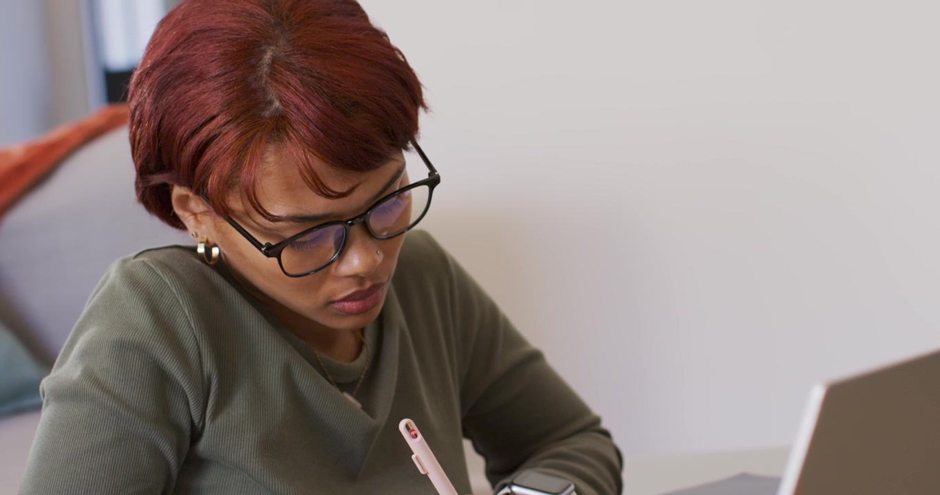 Donna afroamericana che prende appunti alla scrivania per lavoro, controlla l'orologio dopo una notifica, digita sul laptop