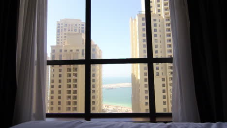 morning light fills a hotel room as a man stands by the window, gazing out at towering buildings and a tranquil sea in the distance