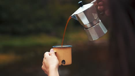 close up shot of a woman pouring hot coffee in a rugged cup out in the forest