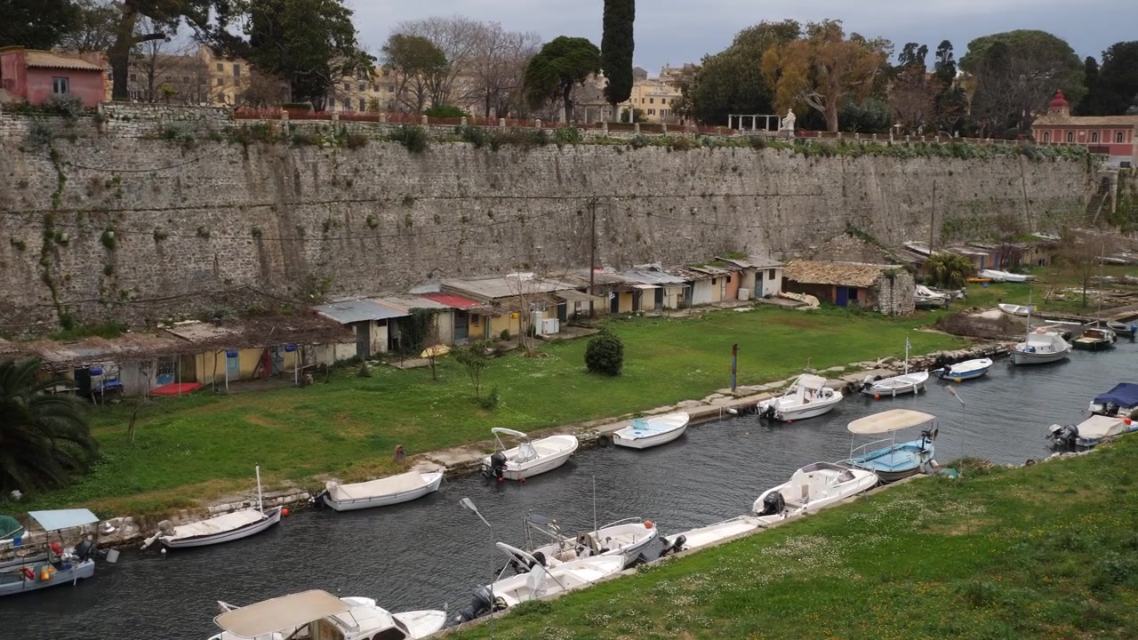 Inquadratura statica dall'alto di piccole imbarcazioni a motore ormeggiate lungo uno stretto canale d'acqua all'interno del fossato di una storica fortezza in pietra, con capanni da pesca sullo sfondo, nella Città Vecchia di Corfù, Isole Ionie, Grecia.