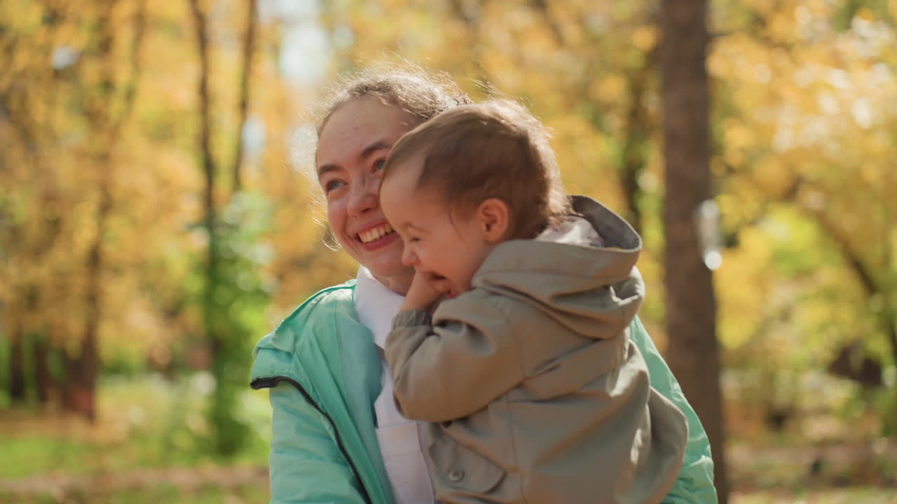 Junge Betreuerin trägt Kleinkind durch Park, Blätter treiben, spontanes Lachen, spielerisches Kitzeln, Schichten von Jacken, sonniges Bokeh, Familienverbindung, unbeschwerter Nachmittagsspaziergang
